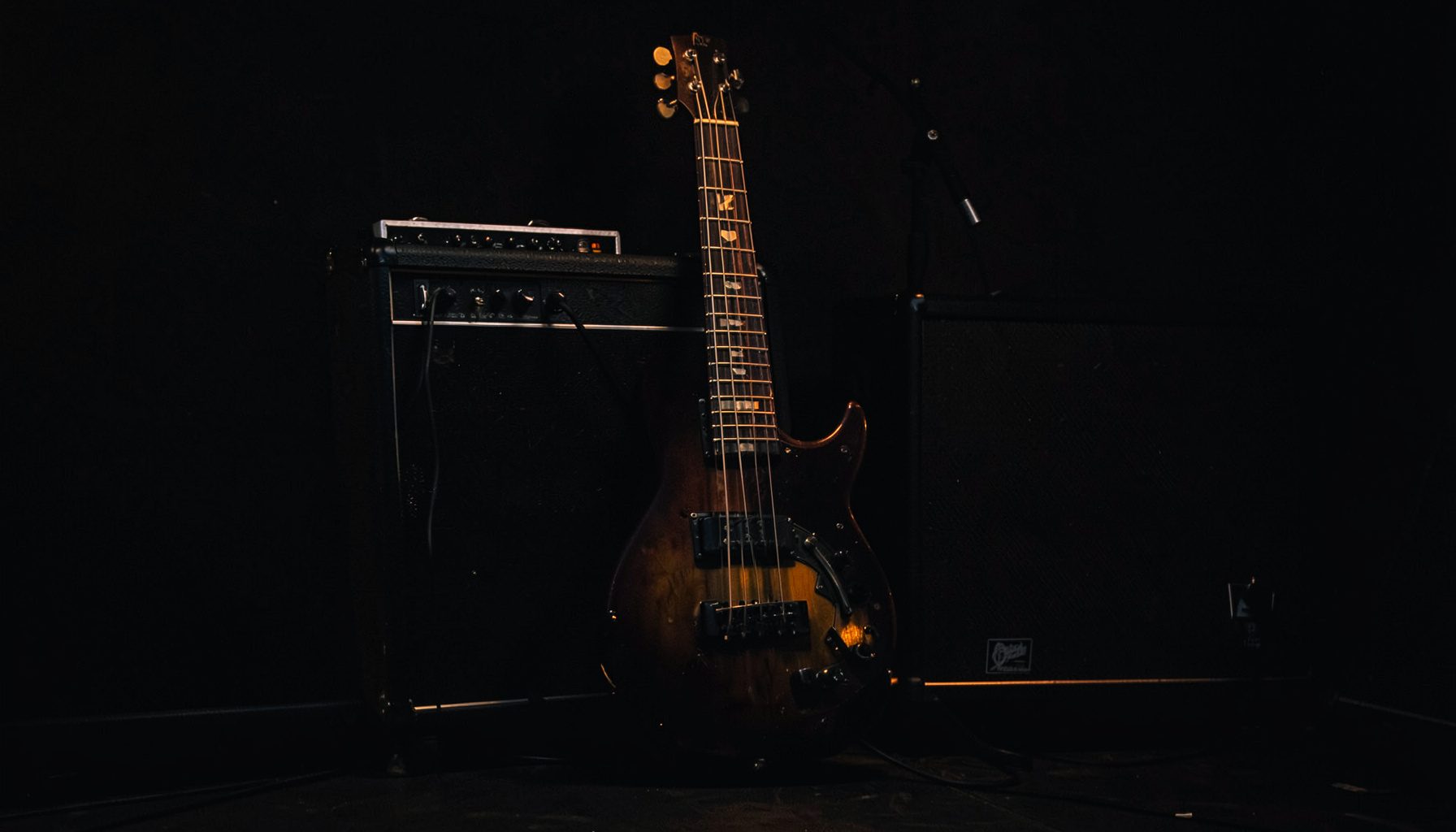 Dark, atmospheric photograph of a bass guitar leaning against a stage amplifier, deep charcoal tones with a subtle amber glow