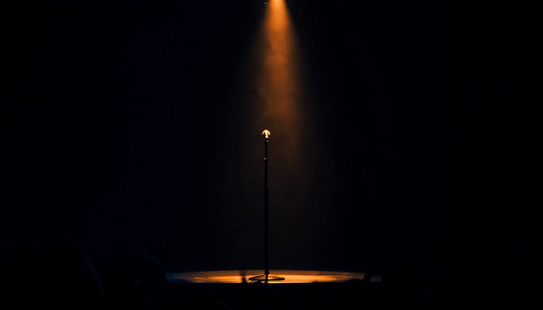 Dark atmospheric photograph of a dimly lit concert stage with deep navy and charcoal shadows, a single warm amber spotlight cutting through the darkness