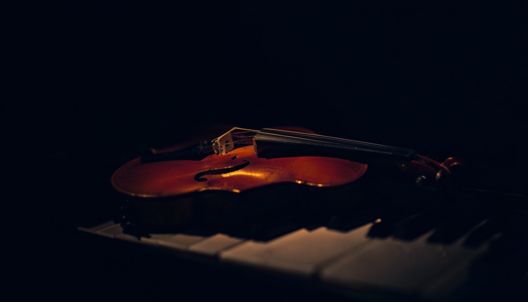 Dark, moody photograph of a violin resting on a piano keyboard, lit by a single soft amber glow against deep navy shadow