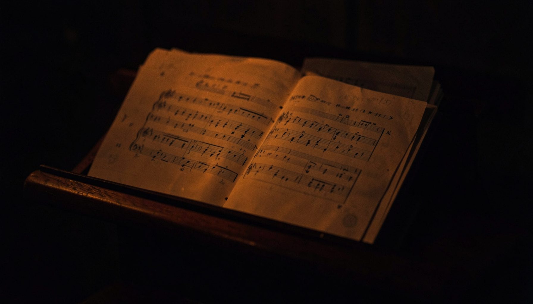 A dark, moody photograph of sheet music resting on a wooden music stand, deep brown and charcoal tones with soft amber light.