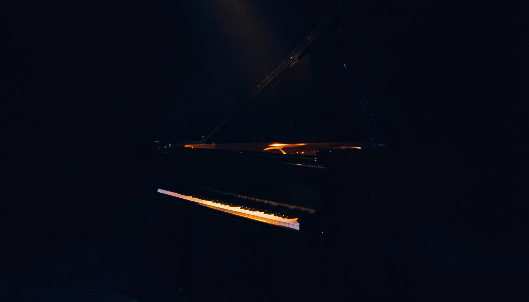 A dark, low-key photograph of a grand piano on a dimly lit stage, deep navy and charcoal shadows with a single warm amber spotlight