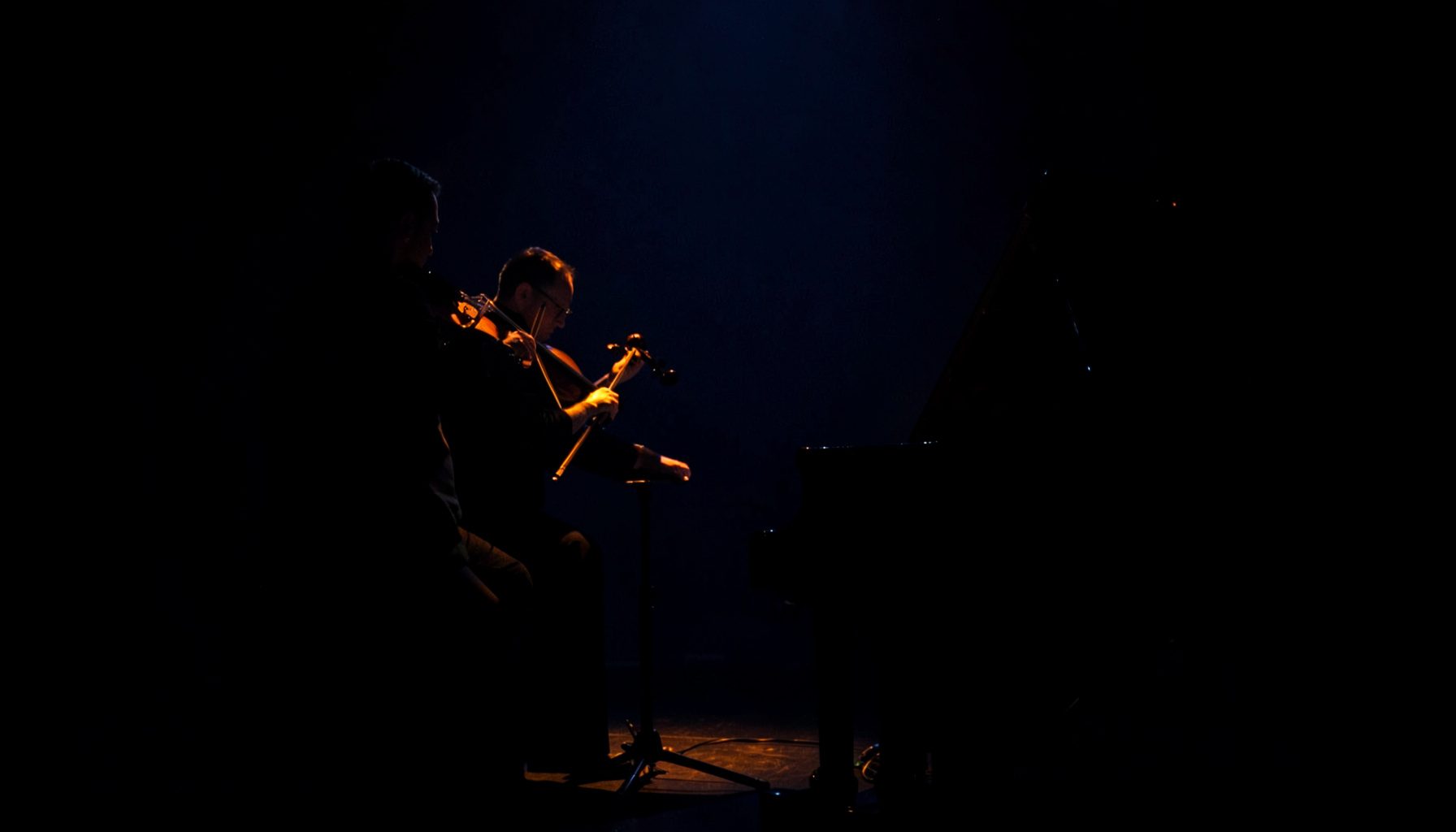 Dark intimate duo performance photograph with violin and piano silhouettes against deep navy and warm amber stage lighting