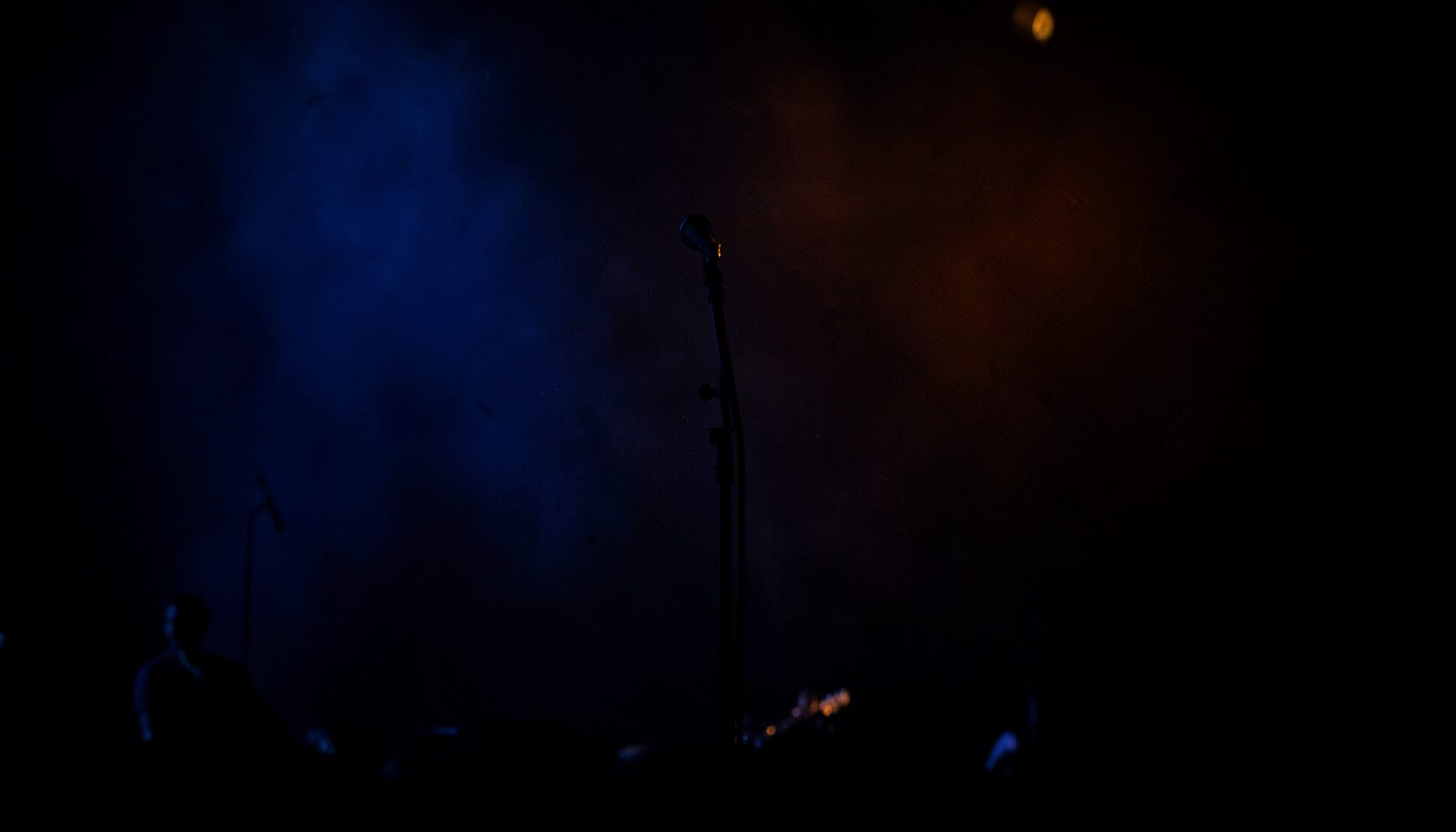 Dark atmospheric photograph of a concert stage with deep blue and amber lighting, empty microphone stand silhouetted against the glow