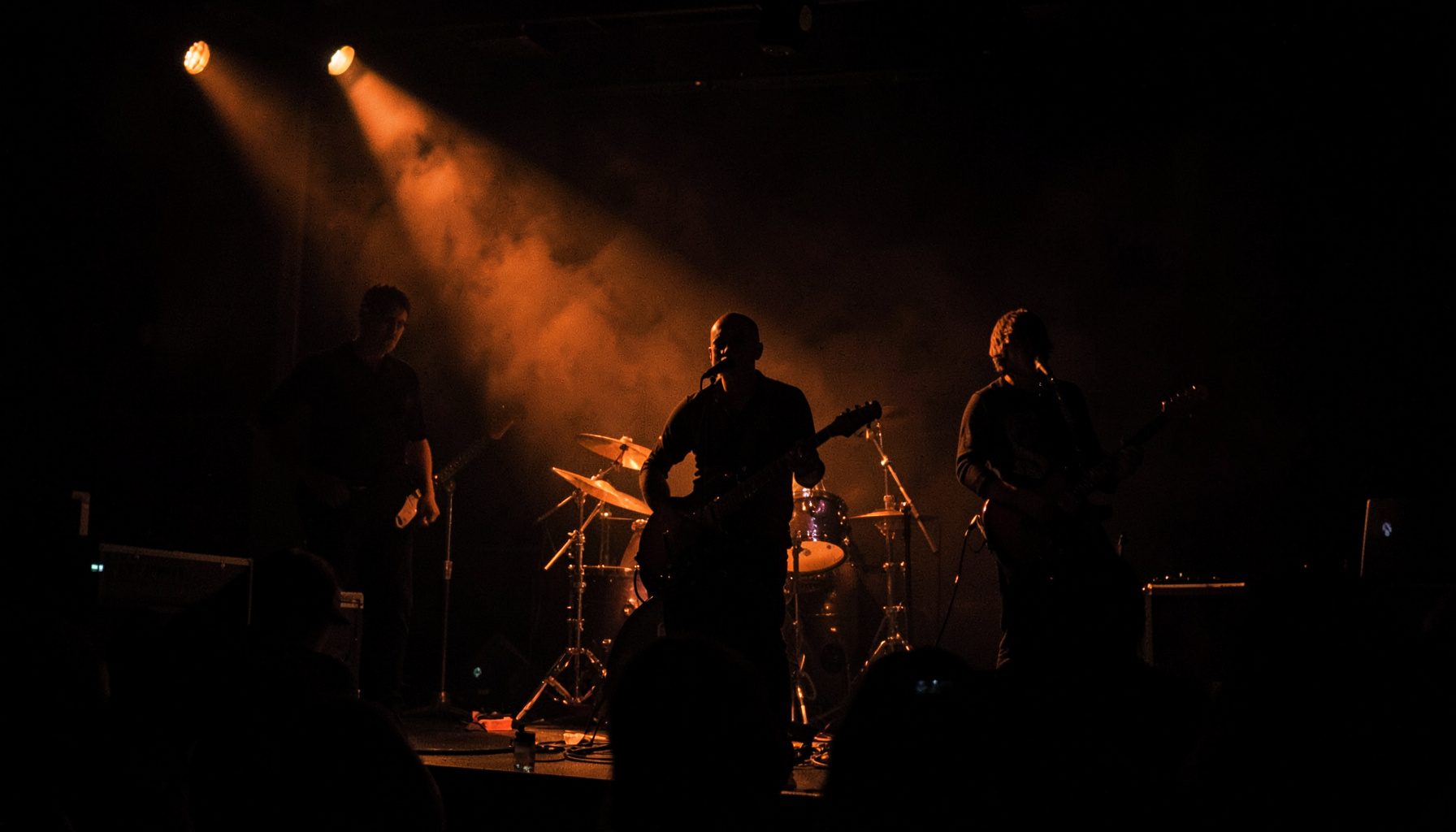 Dark photograph of a rock band performing live, deep shadows with warm amber spotlights cutting through haze on stage