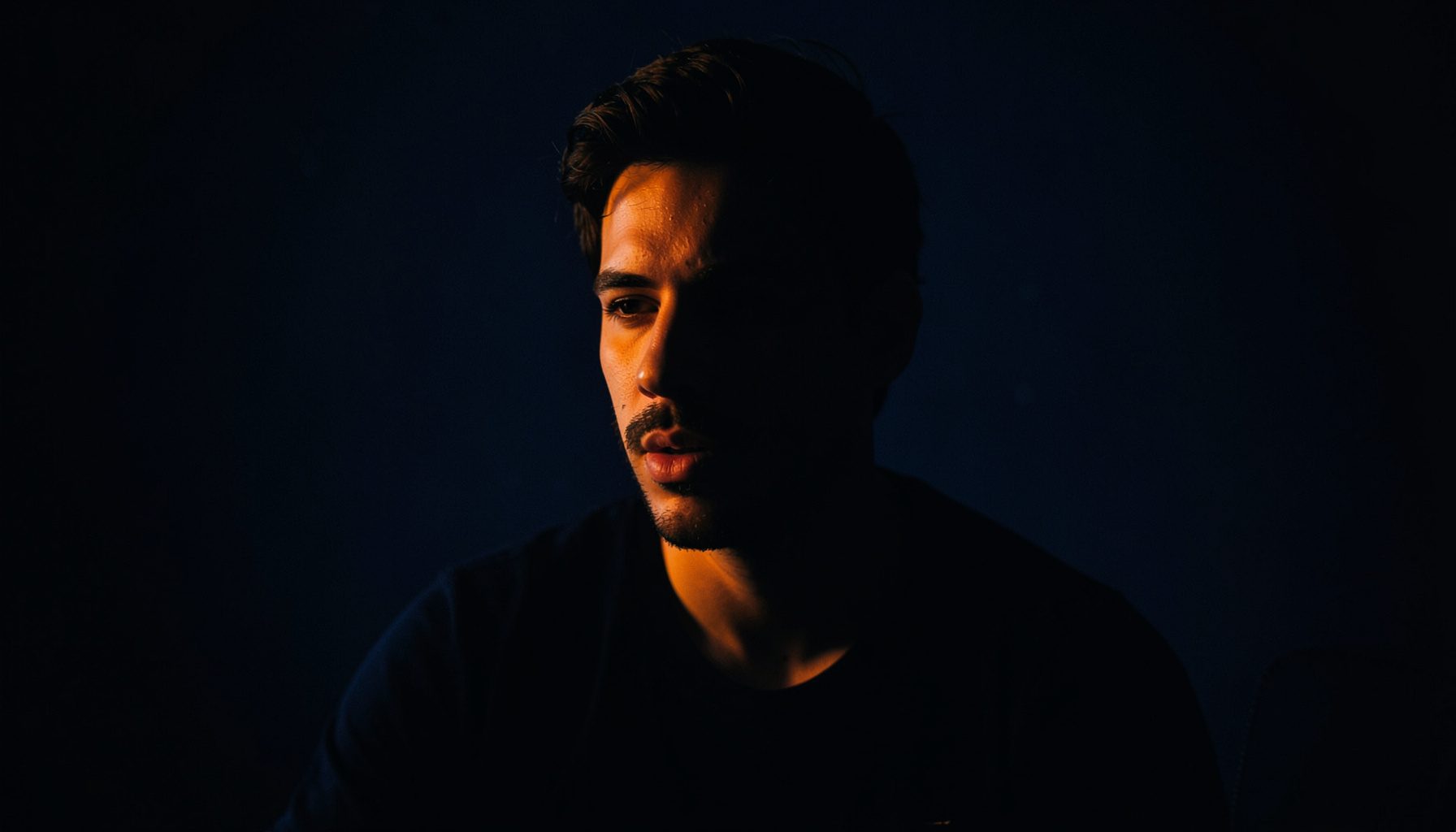 Dark low-key portrait-style photograph of a singer-songwriter in a studio setting, navy tones with a single warm light source