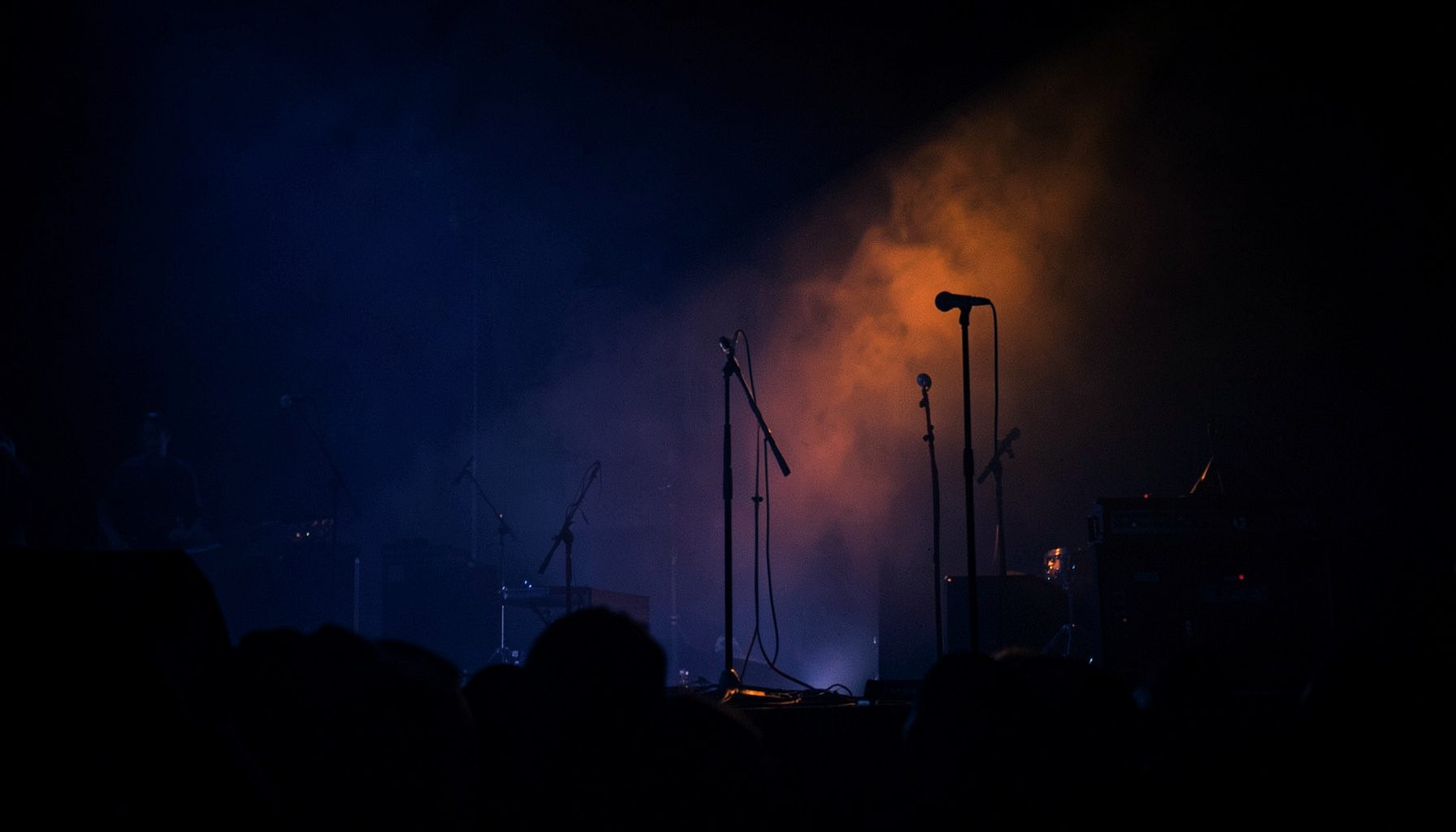 Dark atmospheric photograph of a concert stage with deep navy and amber lighting, microphone stands silhouetted against haze